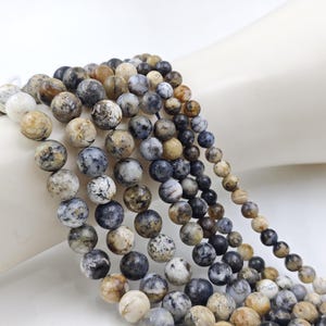 May include: A strand of round, multi-colored gemstone beads. The beads are a mix of brown, white, and gray with a speckled pattern. The beads are strung on a clear thread.