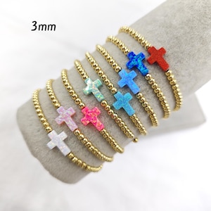 May include: A set of 7 gold beaded bracelets with a small opal cross charm in different colors, including white, pink, blue, and red. The bracelets are 3mm in diameter.