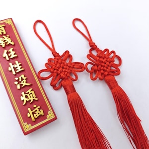 Puede incluir: Dos borlas rojas con nudos chinos con largas franjas y una placa rectangular roja con letras doradas. La placa tiene letras doradas. Las borlas son un símbolo de buena suerte y prosperidad.