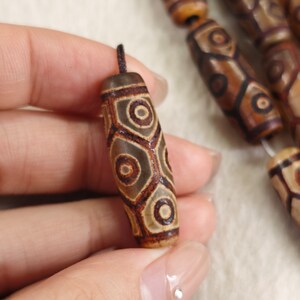 Peut inclure: Gros plan sur une perle d'agate brune et blanche avec un motif hexagonal. La perle est enfilée sur un cordon noir et est tenue par une main.