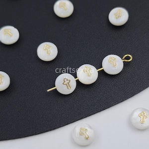 Peut inclure: Perles rondes blanches avec un emblème de croix de couleur dorée centré sur chaque pièce. Trois des perles sont enfilées ensemble sur un fin fil métallique de couleur dorée, tandis que plusieurs autres perles individuelles sont éparpillées.
