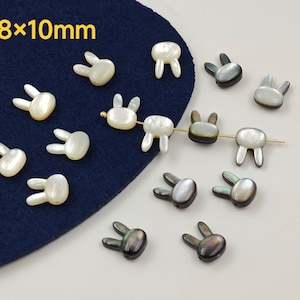 Op de afbeelding: Konijnvormige kralen in wit en iriserend zwart, ongeveer 8 mm x 10 mm groot. Sommige zijn geregen op een gouden draad. De achtergrond bestaat uit donkerblauwe stof en een lichtblauw oppervlak. De tekst "8x10mm" is zichtbaar.