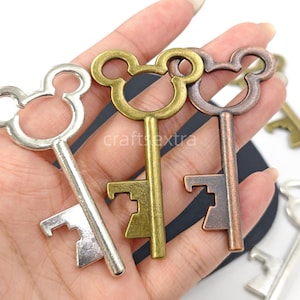 May include: Three metal key-shaped bottle openers in silver, antique gold, and copper tones. Each opener features a circular head with two smaller circular protrusions resembling ears. The shafts are straight with a notched bit at the bottom.