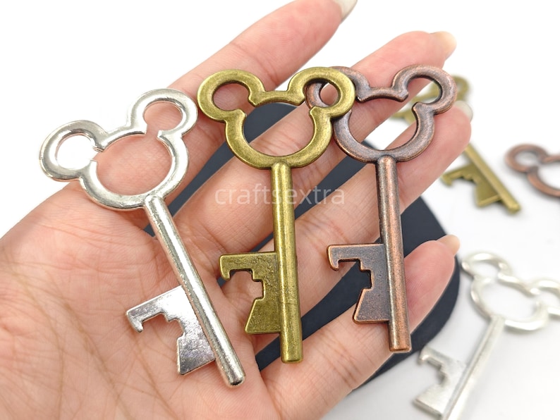 May include: Three metal key-shaped bottle openers in silver, antique gold, and copper tones. Each opener features a circular head with two smaller circular protrusions resembling ears. The shafts are straight with a notched bit at the bottom.