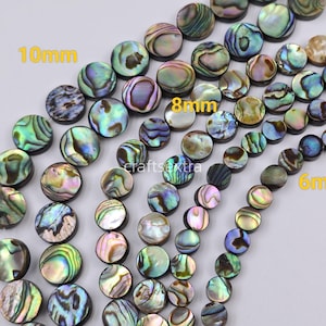 May include: Round abalone shell beads in varying sizes of 10mm, 8mm, and 6mm. The beads feature iridescent, multi-colored surfaces with swirling patterns of green, blue, purple, and gold. The beads are arranged in curved rows on a plain surface.
