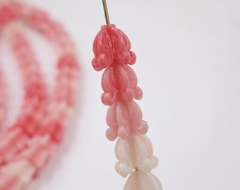 May include: Close-up of a strand of pink and white flower-shaped beads. The beads are strung on a thin gold wire, with a gradient of color from deep pink to white. The background is a soft white.