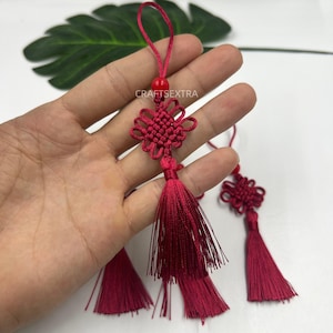 Peut inclure: Un ornement décoratif à pompon rouge foncé avec un nœud complexe tissé, une perle lisse au sommet et une longue frange fluide. L'ornement comprend une boucle pour suspendre et le texte CRAFTSEXTRA est visible au centre de l'objet.