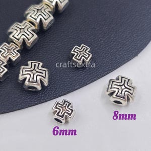 May include: Silver-toned metal beads shaped like crosses with an embossed geometric pattern. The beads are shown in two sizes, labelled as 6mm and 8mm, with a central hole for threading. The text craftsextra is visible in the centre of the image.