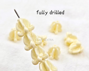May include: A collection of off-white, flower-shaped beads with a glossy finish. Several beads are strung on a thin metal wire, while others are scattered nearby. The text 'fully drilled' and 'CRAFTSEXTRA' are visible in the image.