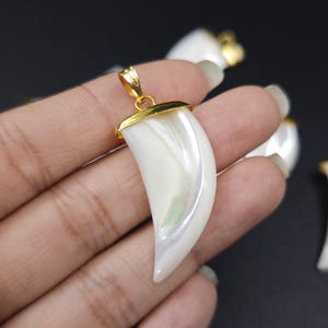 Puede incluir: Varios colgantes nacarados en forma de garra, con detalles metálicos dorados. Los colgantes blancos tienen diseños iridiscentes y se muestran sobre un fondo oscuro.