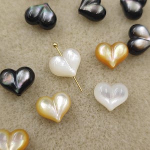Pode incluir: Contas iridescentes em forma de coração em várias cores, incluindo preto, branco e dourado. Um alfinete dourado é inserido em uma das contas brancas. As contas estão espalhadas sobre uma superfície castanha clara. Estas contas são adequadas para fazer joias.