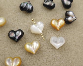 May include: Heart-shaped iridescent beads in various colors, including black, white, and gold. A gold-colored pin is inserted into one of the white beads. The beads are scattered on a light brown surface. These beads are suitable for jewelry making.