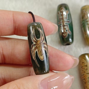 Puede incluir: Un conjunto de cinco cuentas de ágata verde con diseños de arañas marrones y blancas. Las cuentas están enhebradas en un cordón negro.