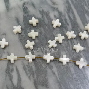 May include: A collection of small, white, cross-shaped beads made from mother of pearl. Several of these beads are loose, while five are strung together on a thin, brown cord. The beads have a smooth, slightly iridescent surface finish.