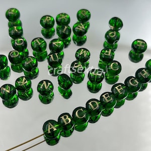 Peut inclure: Une collection de perles rondes en verre vert foncé translucide, chacune portant une lettre de l'alphabet en couleur dorée. Un fil est disposé dans l'ordre alphabétique de A à I, tandis que les autres perles sont éparpillées autour.