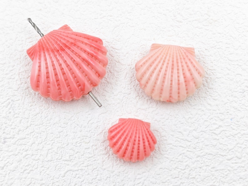 K&ouml;nnte beinhalten: Drei rosa Muscheln mit einem gebohrten Loch oben. Die Muscheln sind unterschiedlich gro&szlig; und haben eine strukturierte Oberfl&auml;che.