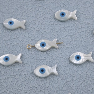 Może przedstawiać: Białe koraliki w kształcie rybki z niebieskim wzorem oka. Koraliki mają perłowe wykończenie i złoty uchwyt do nawlekania. Te ozdobne koraliki są idealne do tworzenia biżuterii lub projektów rzemieślniczych.