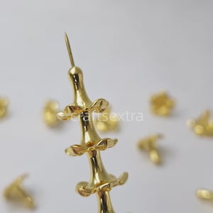 Puede incluir: Una colección de chinchetas de metal de color dorado, cada una con una punta afilada y un eje decorativo escalonado similar al bambú con tres bridas circulares distintivas en forma de hoja. Las chinchetas están dispuestas con una enfocada.