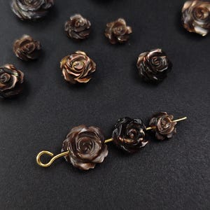 May include: A collection of dark brown rose-shaped beads and components. Several individual beads are scattered across a dark surface. A group of three beads are strung together on a gold-colored wire, with a loop at one end. The beads have a detailed, carved appearance.