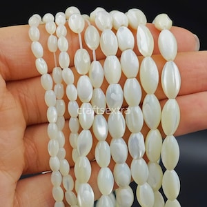 Peut inclure: Plusieurs rangées verticales de perles en nacre blanche de forme ovale. Les perles présentent une finition lisse et irisée et sont enfilées sur un fil transparent fin. Le texte "craftextra" est visible au centre de l'image.