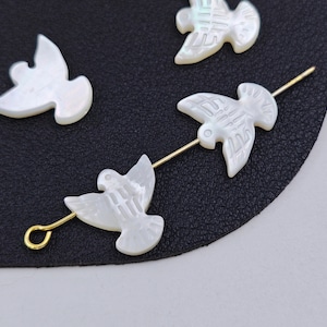 May include: Four white mother-of-pearl dove charms with gold-colored wire attachments. The charms are carved with detailed wing and body patterns. The doves are arranged on a black surface, with one dove attached to a gold wire loop.