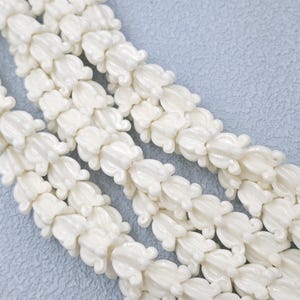 May include: Close-up of several strands of white, flower-shaped beads. The beads are strung together, creating a continuous pattern. The beads have a glossy finish and are arranged diagonally across the frame.