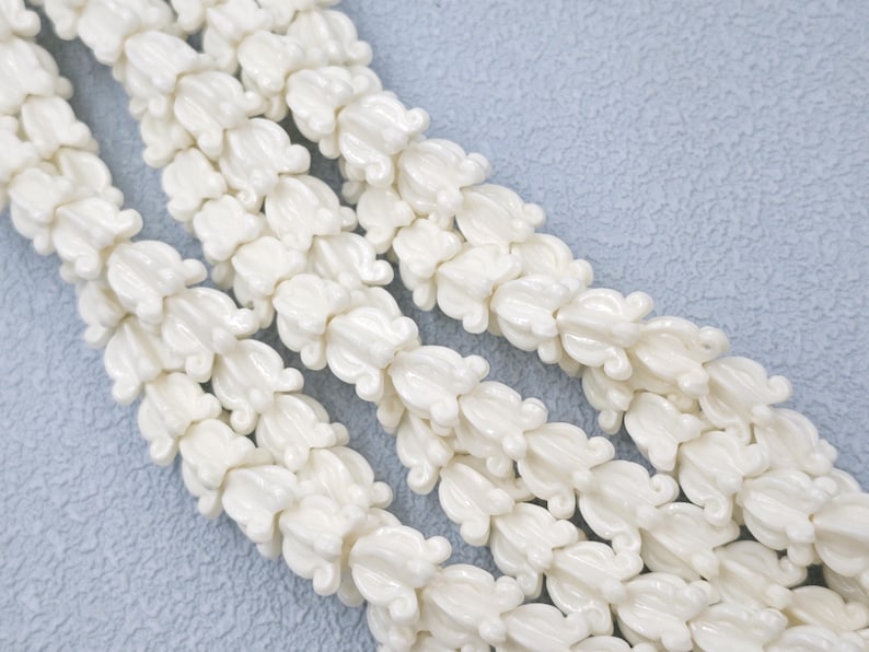 May include: Close-up of several strands of white, flower-shaped beads. The beads are strung together, creating a continuous pattern. The beads have a glossy finish and are arranged diagonally across the frame.