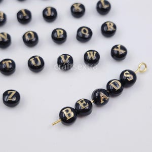 Könnte beinhalten: Eine Sammlung runder schwarzer Perlen mit goldfarbenen Buchstaben des Alphabets. Ein goldfarbener Metalldraht hält fünf Perlen, die das Wort BEADS bilden, mit einer kleinen Schlaufe an einem Ende. Die restlichen Perlen liegen verstreut daneben.