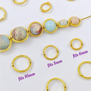 Könnte beinhalten: Goldfarbene Sprungringe in verschiedenen Größen, mit dem Text "fits 6mm", "fits 8mm" und "fits 10mm" darunter gedruckt. Die Ringe sind um eine Reihe von bunten Edelsteinperlen angeordnet.