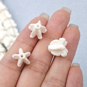 May include: Three small, off-white, star-shaped and flower-shaped beads. The beads have a hole in the center for stringing. The beads are held in a hand against a light blue background. The beads are likely for jewelry making.
