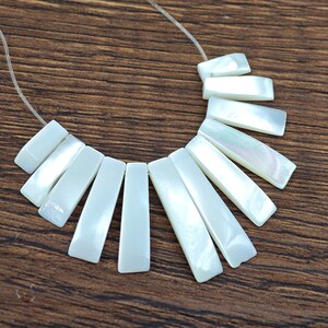 May include: A white mother-of-pearl necklace with a fan-shaped design. The necklace is made of rectangular pieces of shell that are strung together on a clear cord.