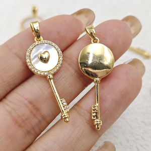 May include: Two gold key-shaped pendants with a round charm. One charm is a white mother-of-pearl with a gold heart and clear rhinestones around the edge. The other charm is solid gold and shiny.