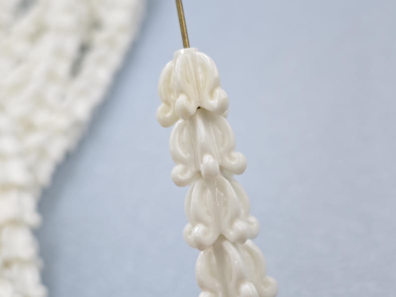May include: Close-up of a strand of white, flower-shaped beads on a gold pin. The beads are clustered together, with a delicate, textured appearance. The background is a soft, light blue.