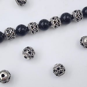 May include: A strand of black beads with silver filigree accents. The beads are round and have a polished finish. Several loose beads are scattered around the strand on a white surface. These beads could be used for jewelry making.