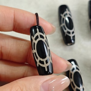 Peut inclure: Pendentif noir de forme oblongue avec un motif géométrique clair. Le pendentif est attaché à un cordon foncé en haut. Plusieurs pendentifs similaires sont visibles en arrière-plan.