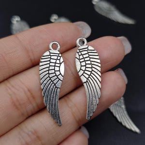 Könnte beinhalten: Zwei silberne Engelsflügel-Anhänger mit detailliertem Design. Die Flügel sind oben an einer Schlaufe befestigt, um sie leicht an Schmuck oder Bastelarbeiten anzubringen.