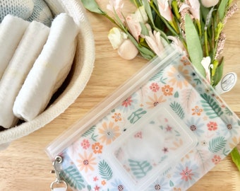 May include: A floral patterned baby wipes case with a clear plastic lid and a white strap. The case is lying on a wooden surface next to a basket of white items and a bouquet of flowers.