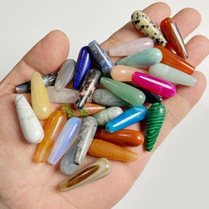 May include: A collection of colorful gemstone pendants in various shapes and sizes. The pendants are in shades of blue, green, pink, orange, and white. Some have unique patterns and textures, while others are smooth and polished. The pendants are held in a hand.