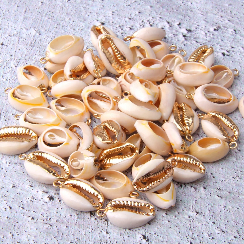 Puede incluir: Una colecci&oacute;n de conchas de cauri blancas con detalles dorados. Las conchas tienen un agujero perforado y est&aacute;n listas para ser usadas para la elaboraci&oacute;n de joyas o manualidades.
