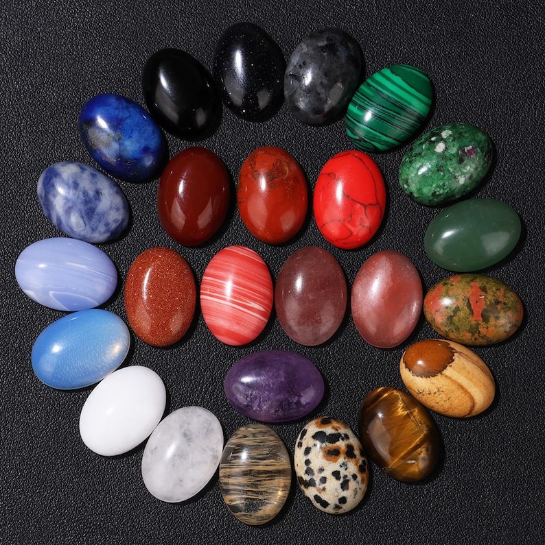 K&ouml;nnte beinhalten: Eine Sammlung von einundzwanzig ovalf&ouml;rmigen Edelsteinen in verschiedenen Farben, darunter Blau, Rot, Gr&uuml;n und Braun. Die Steine sind in einem kreisf&ouml;rmigen Muster auf einer schwarzen Oberfl&auml;che angeordnet und zeigen ihre polierten Oberfl&auml;chen und nat&uuml;rlichen Muster.