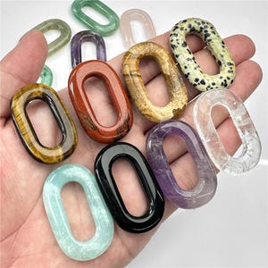 May include: Assortment of oval-shaped gemstone links in various colors, including green, pink, brown, red, white, black, and purple. The links are smooth and polished, with a hole in the center for attaching to a chain or cord.