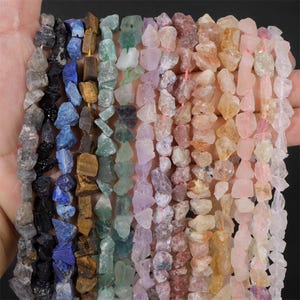 May include: A collection of gemstone chip beads in a variety of colors, including black, blue, green, purple, and orange. The beads are strung together on a pink string, creating a colorful and textured display. Ideal for jewelry making.