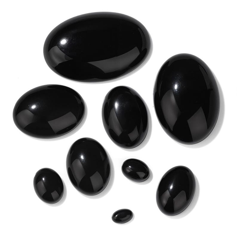 K&ouml;nnte beinhalten: Eine Sammlung polierter, oval geformter, schwarzer Edelsteine in verschiedenen Gr&ouml;&szlig;en. Die Steine haben eine gl&auml;nzende, reflektierende Oberfl&auml;che und sind vor einem wei&szlig;en Hintergrund angeordnet. Die Edelsteine sind wahrscheinlich f&uuml;r Schmuck oder Dekorationszwecke bestimmt.