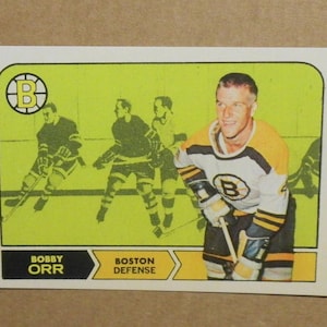 May include: A vintage hockey trading card featuring Bobby Orr, a Boston Bruins player. The card is yellow with black and white accents. The card features a photo of Orr in his Bruins uniform, holding a hockey stick. The card also includes the text "Bobby Orr" and "Boston Defense".