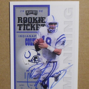 May include: A collectible rookie ticket featuring a football player in a white and blue uniform, holding a football. The ticket includes the text "PLAYOFF 98 ROOKIE TICKET" and the player's signature. The Indianapolis Colts logo is also visible.