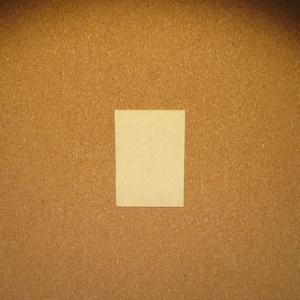 May include: A rectangular, light beige paper card with a smooth, matte surface. The card is positioned centrally against a textured, brown cork board background, creating a clear contrast between the light paper and the dark, granular surface.