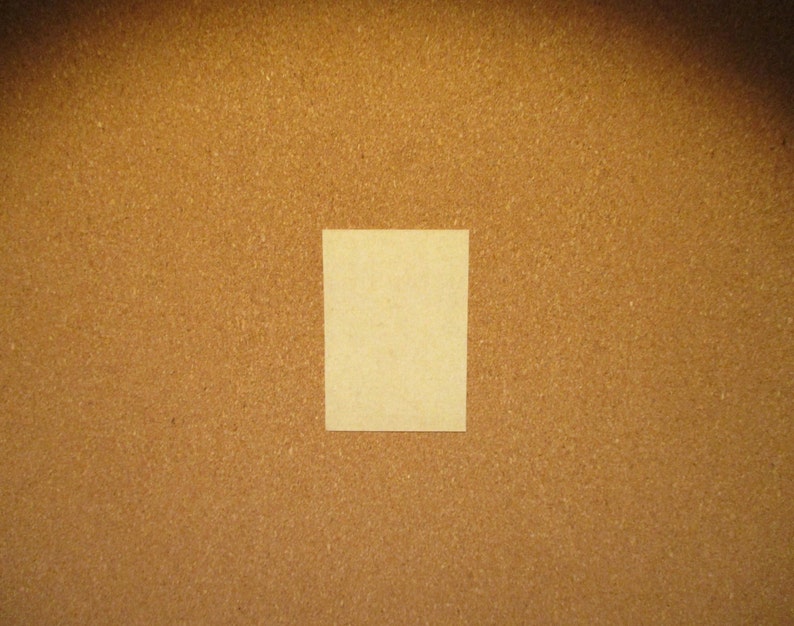 May include: A rectangular, light beige paper card with a smooth, matte surface. The card is positioned centrally against a textured, brown cork board background, creating a clear contrast between the light paper and the dark, granular surface.