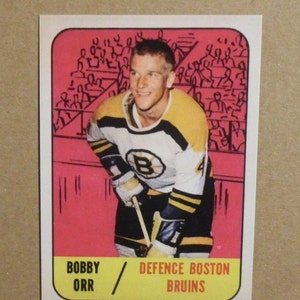 May include: A vintage hockey card featuring Bobby Orr, a defenceman for the Boston Bruins. The card is yellow and features a photo of Orr in his Bruins uniform, holding a hockey stick. The card also includes the text "Bobby Orr / Defence Boston Bruins".