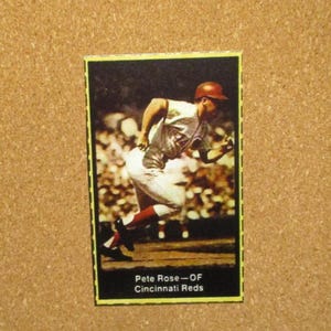 May include: A vintage baseball card featuring Pete Rose, an outfielder for the Cincinnati Reds. The card is yellowed with age and has a black background with white text.