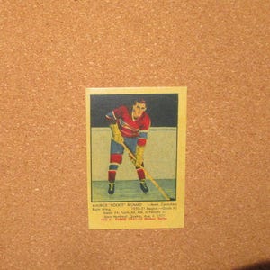 May include: A vintage rectangular hockey trading card featuring an illustration of Maurice Rocket Richard in a red and blue uniform holding a hockey stick. The card includes printed text detailing his 1950-51 record, position, and birth date.